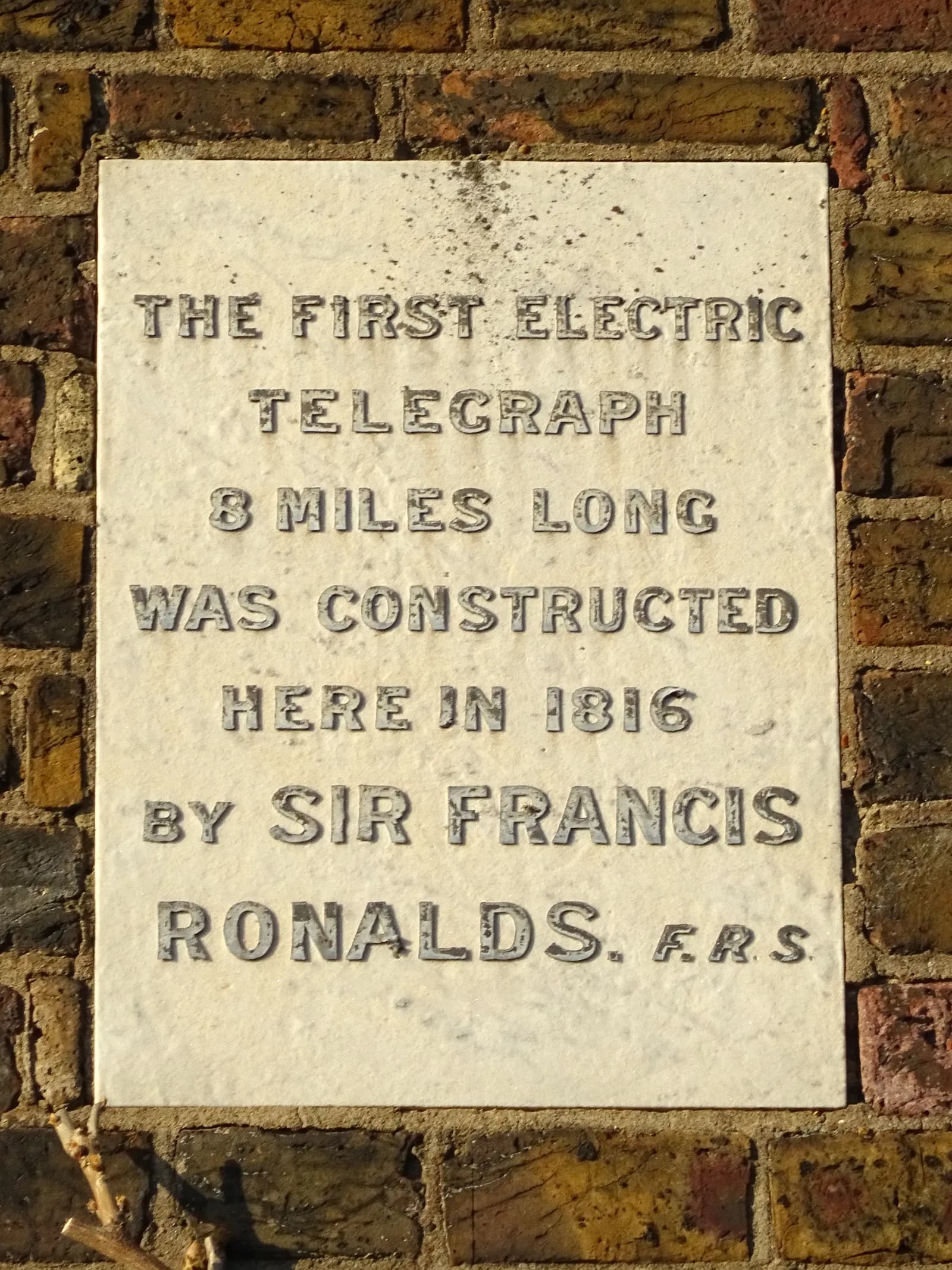 A light-colored plaque on a brick wall commemorating the construction of the first electric telegraph by Sir Francis Ronalds in 1816.