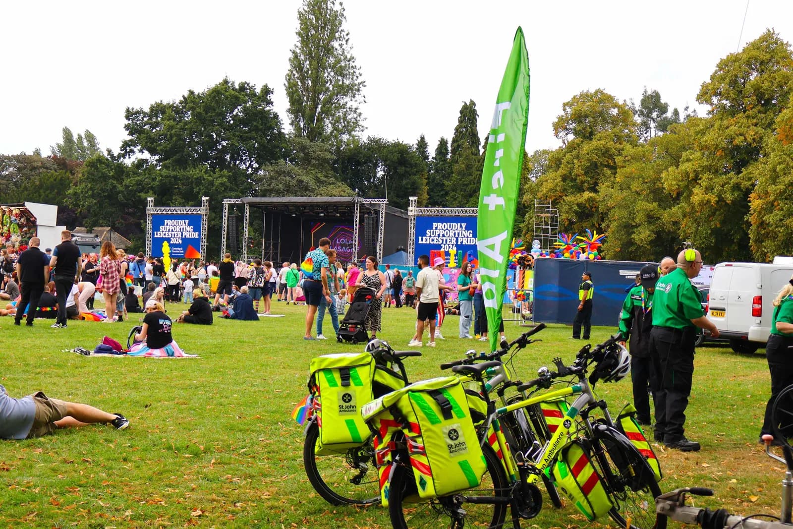 A park scene with people gathered on a grassy area, a stage in the background, and bicycles with bright yellow bags in the foreground, during an outdoor event.
