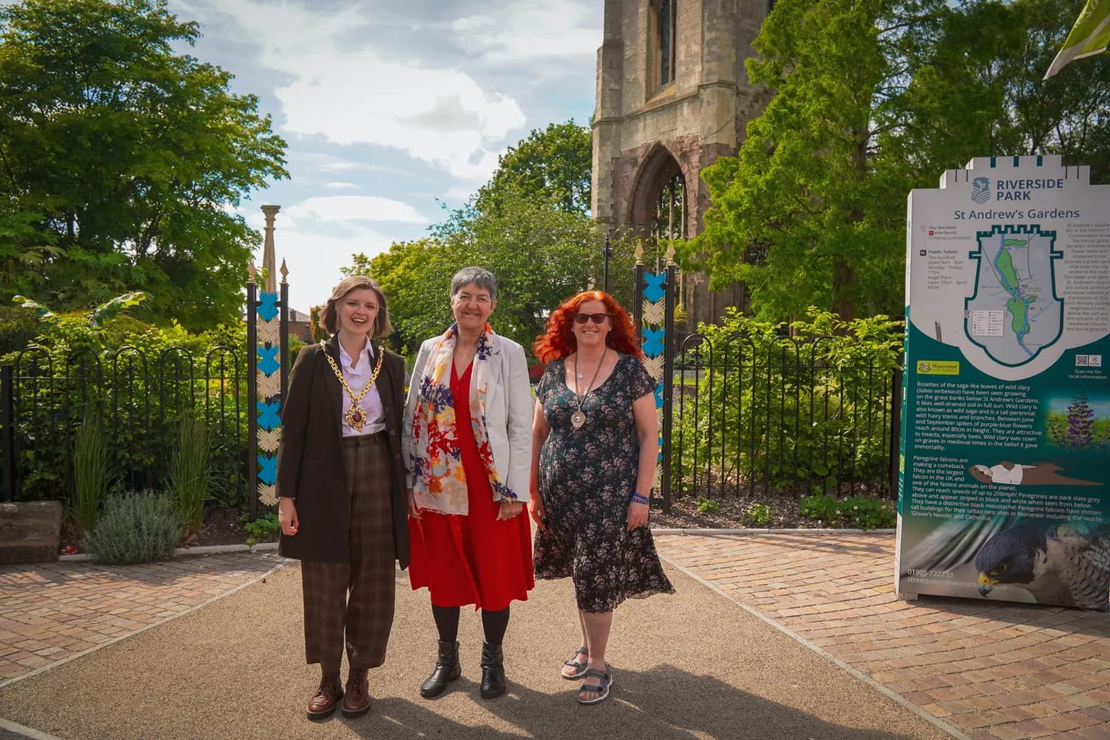 Three women standing next to a park sign on a paved path, with a partial view of an old stone building in the background.