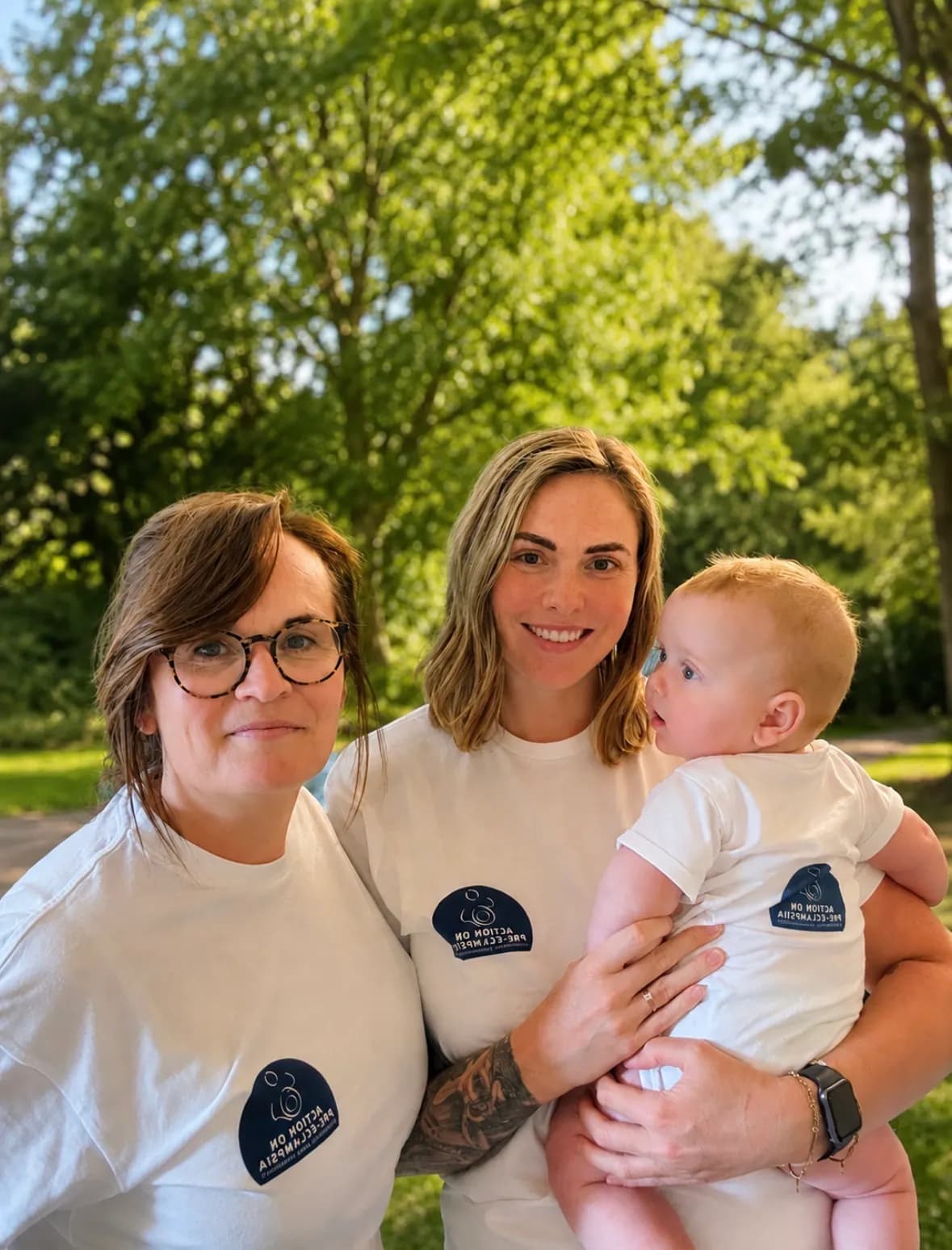 Two women wearing white t-shirts with a blue logo, one holding a baby, stand in front of a leafy green background.