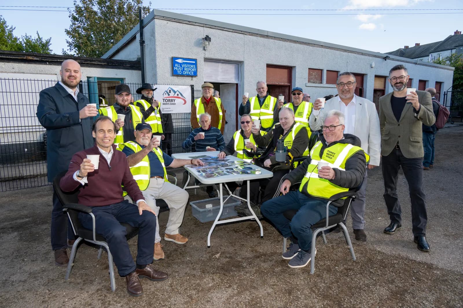 A group of men, some wearing yellow safety vests, are gathered outdoors around a white table, holding cups and smiling at the camera.