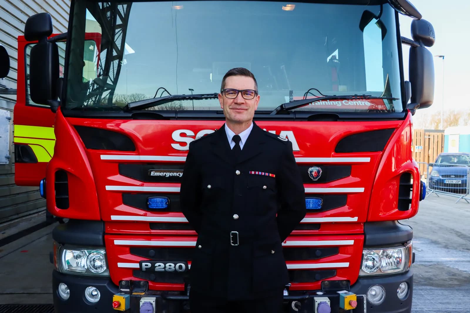 A man in a dark uniform with a tie and glasses smiles at the camera while standing in front of a large red fire truck.