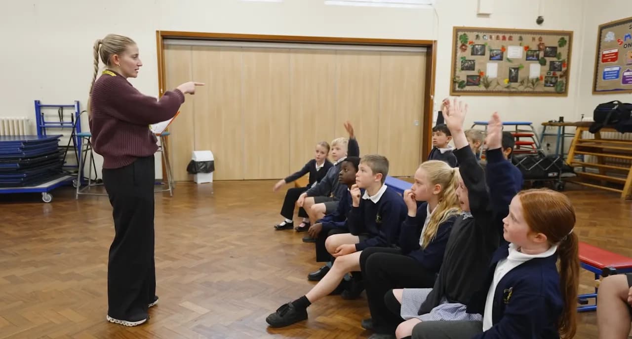 A teacher in a maroon sweater points towards a group of young students sitting on the floor, some raising their hands.