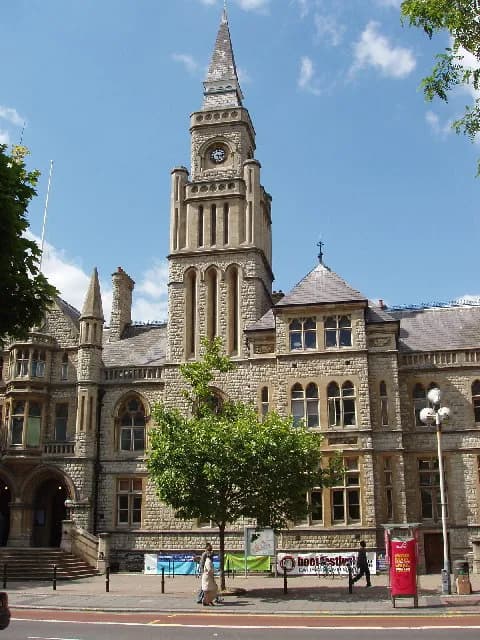 A grand stone building with an ornate clock tower stands tall against a clear blue sky, with a tree in front and people walking on the sidewalk.
