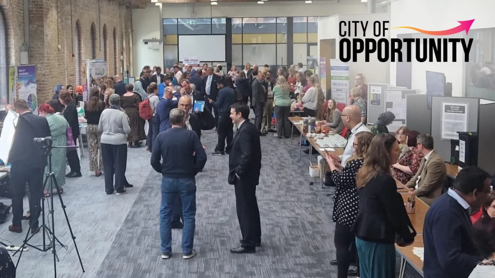 A diverse group of people mingle and network at a business event, with a large “CITY OF OPPORTUNITY” logo visible in the upper right.