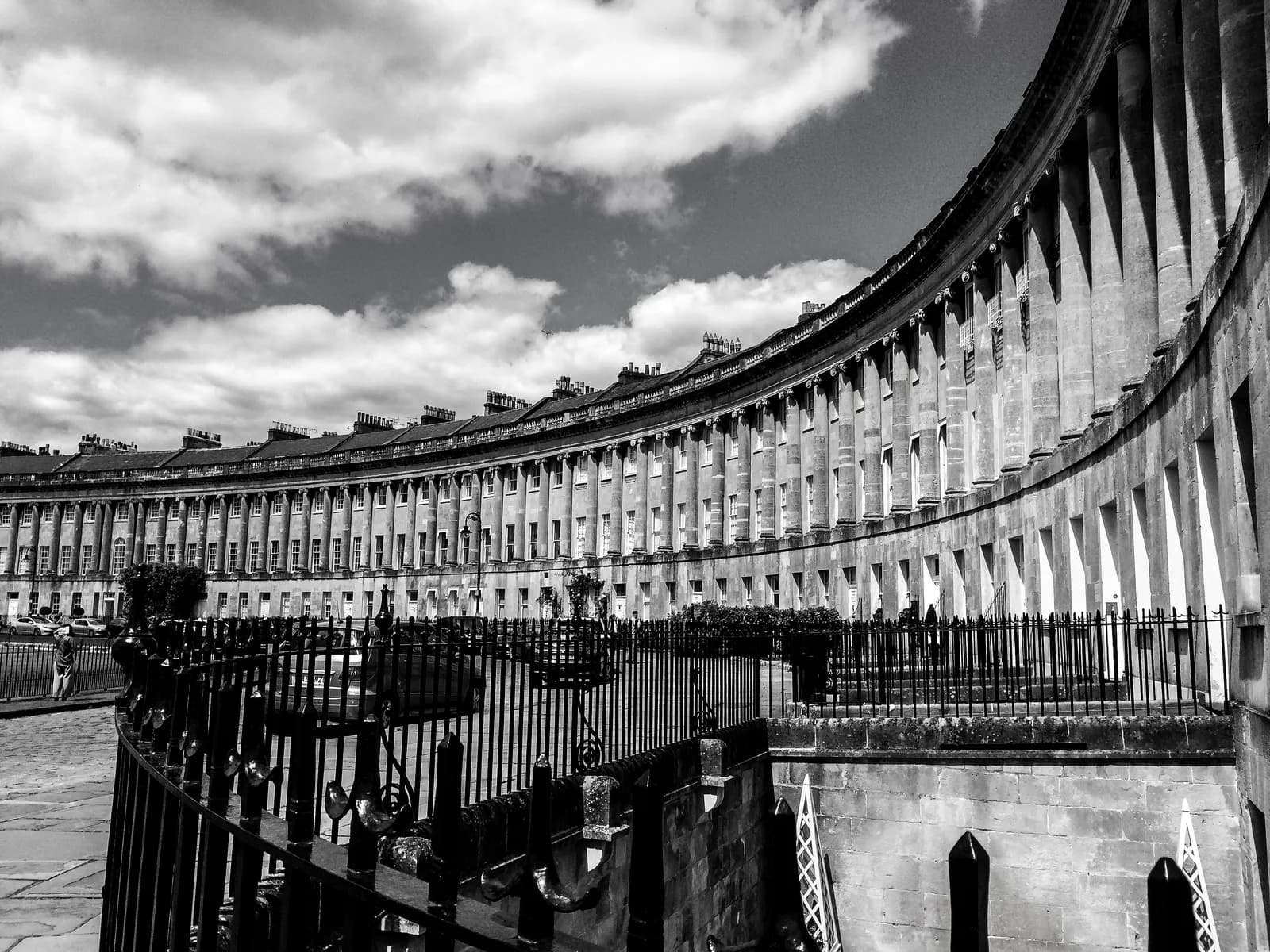 A grayscale photograph shows the Royal Crescent in Bath, England, a curved row of elegant Georgian townhouses with classical columns, under a cloudy sky.