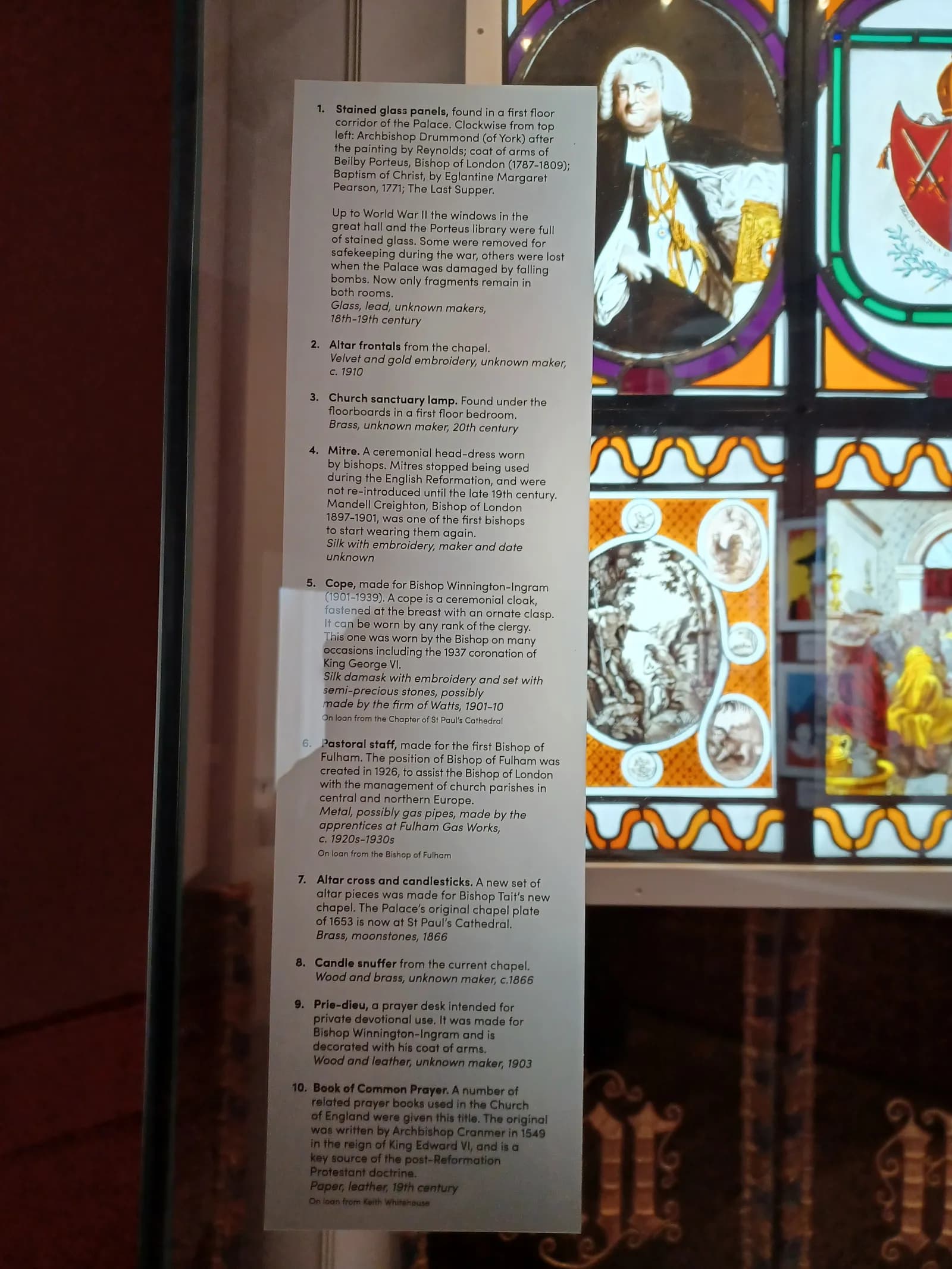 A display case shows information about stained glass panels and various religious artifacts, with portions of the stained glass visible to the right and a reflection of a person in the display case.