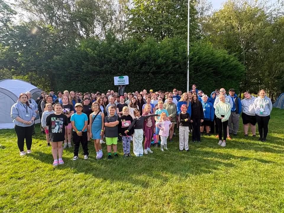 A large group of children and adults pose for a photo outdoors in a grassy area with tents and trees in the background, with some wearing blue hoodies and others in various casual attire.