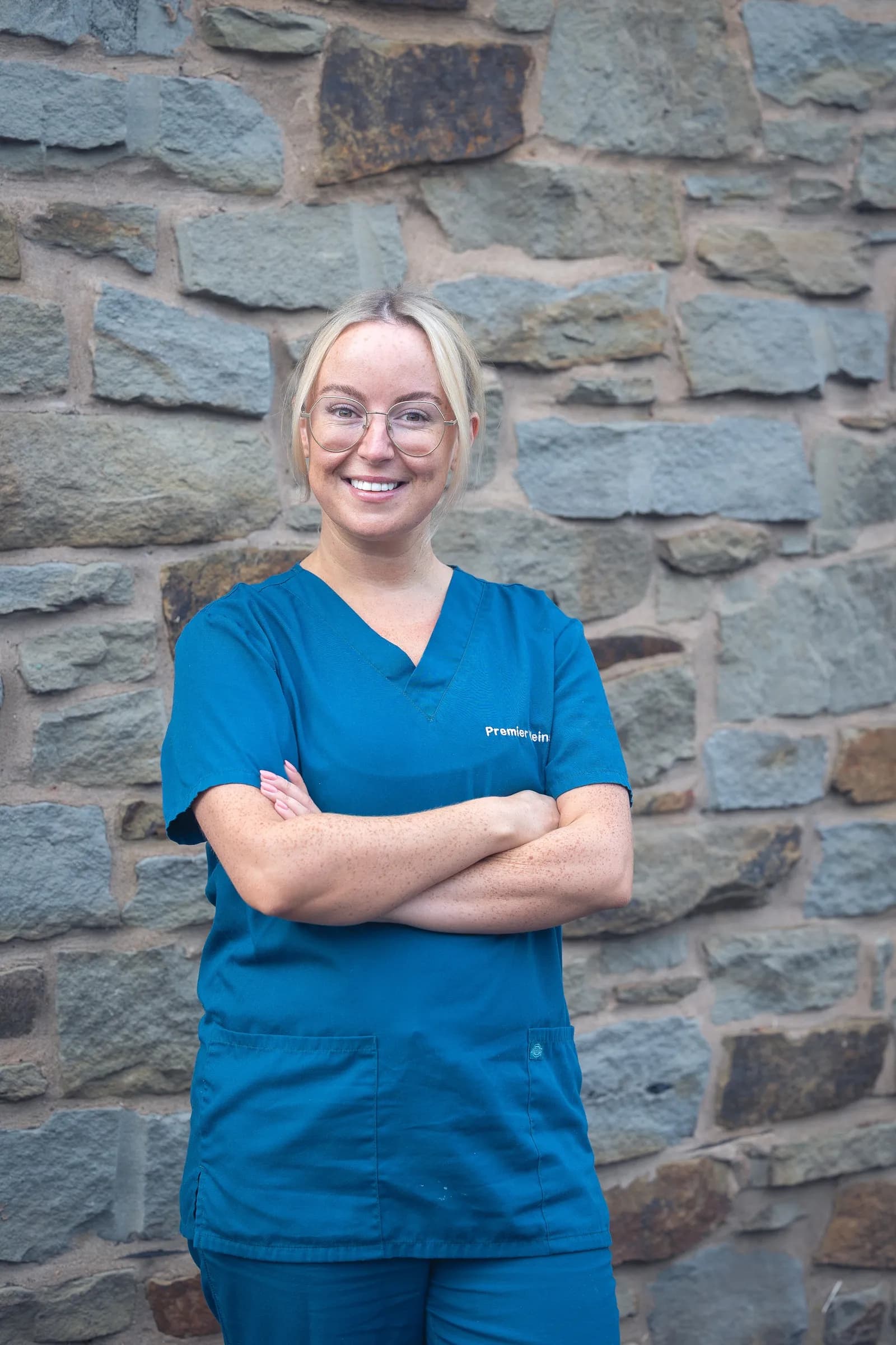 A smiling woman with blonde hair and glasses, wearing a blue medical scrub uniform and standing with her arms crossed in front of a stone wall.