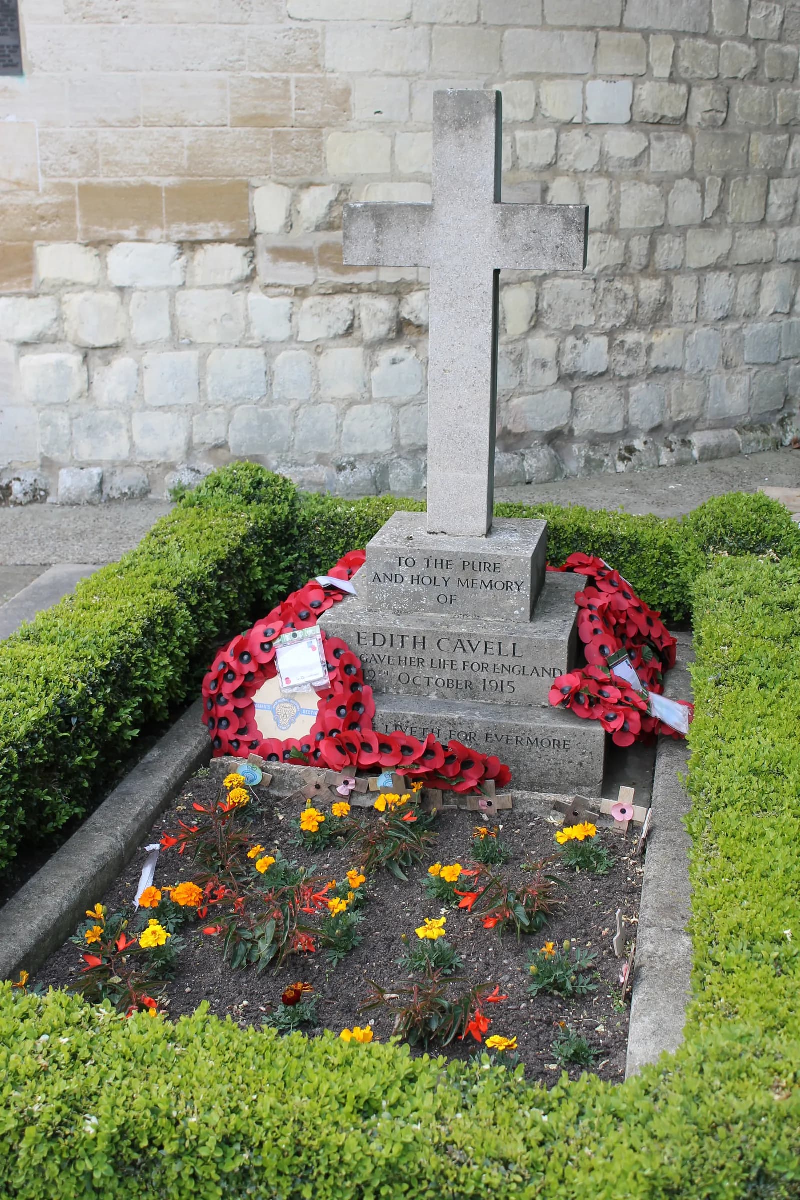 The grave of Edith Cavell with a stone cross headstone and two poppy wreaths, surrounded by green hedges and yellow and orange flowers.