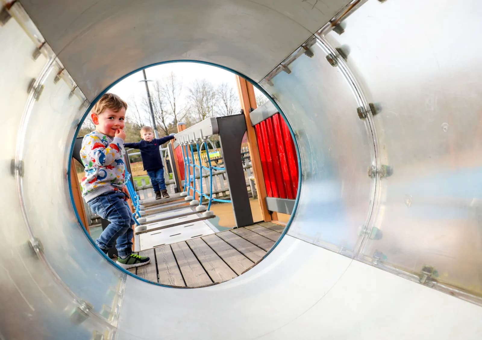 A young boy with a dinosaur sweatshirt and jeans peeks out from the end of a metal tunnel in a playground, looking toward another boy on a wooden bridge.