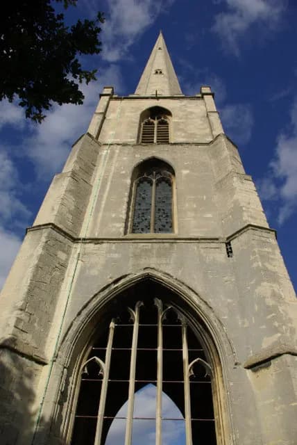 A towering stone church steeple with intricate arched windows reaches towards a bright blue sky with scattered clouds, with dark tree branches visible in the upper left.