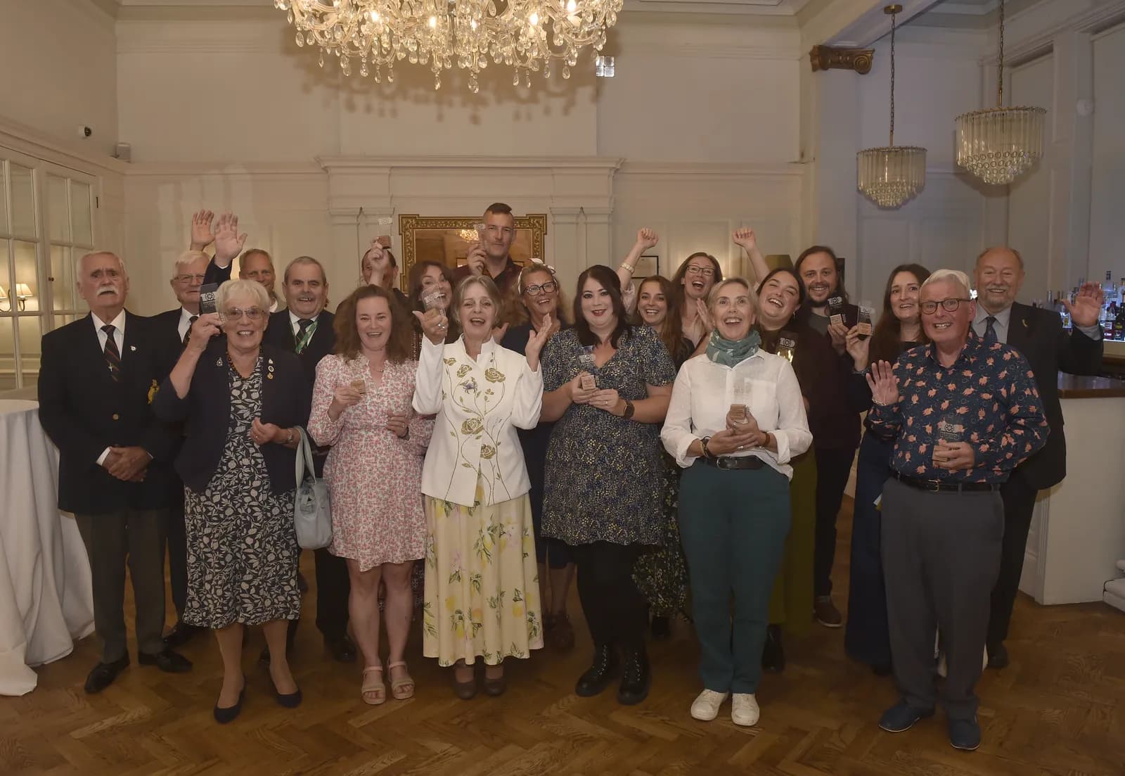 A diverse group of people of all ages stand in a brightly lit room, smiling and holding up glasses in celebration.