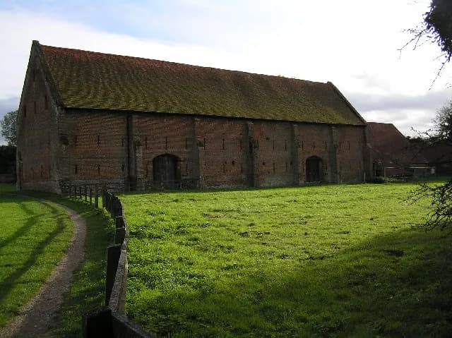 A long brick barn with a mossy brown roof stands in a green field under a partly cloudy sky, with a dirt path and wooden fence in the foreground.