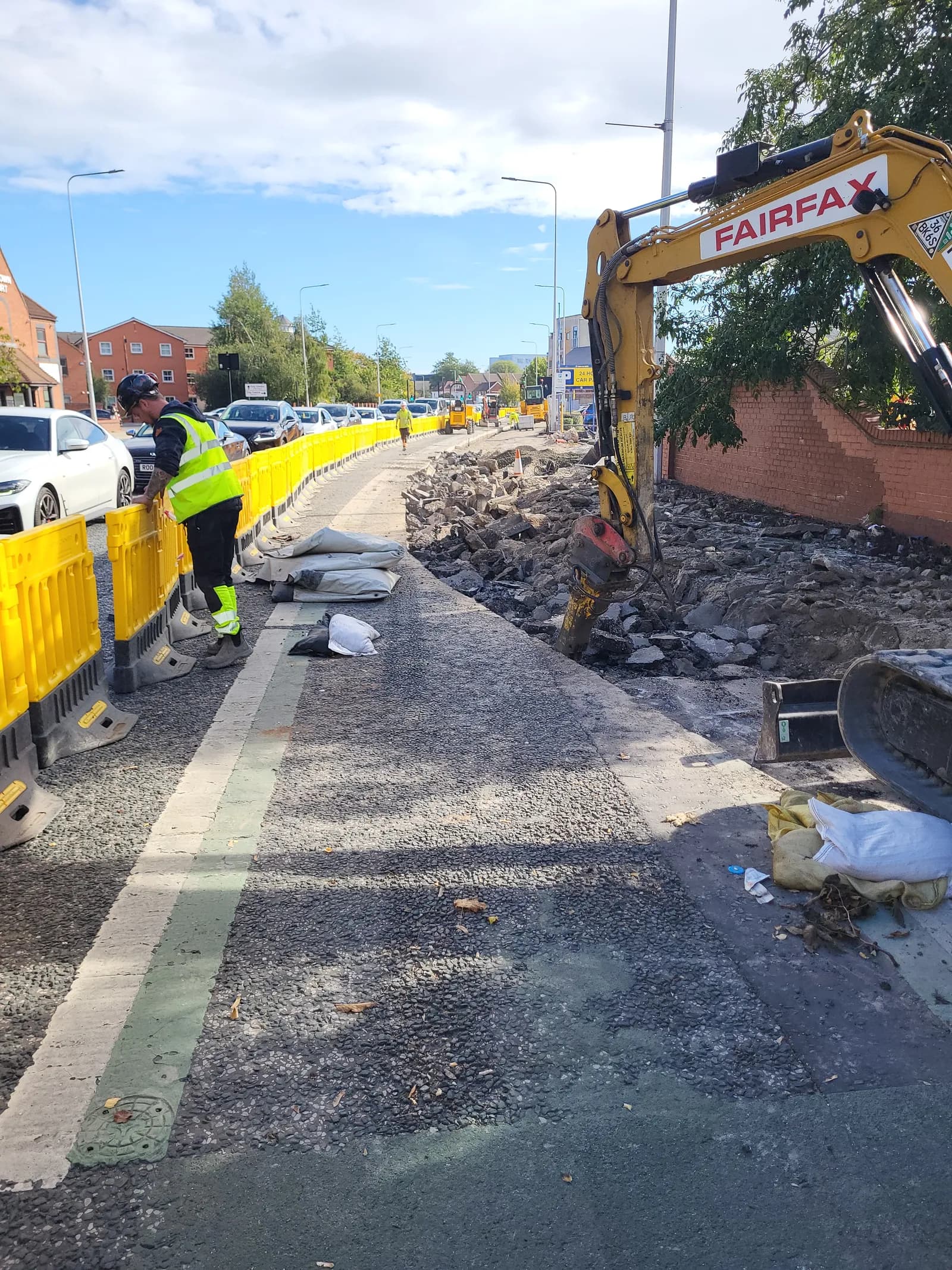A construction worker in a high-visibility vest leans against a yellow barrier next to a construction site with a yellow excavator and debris.