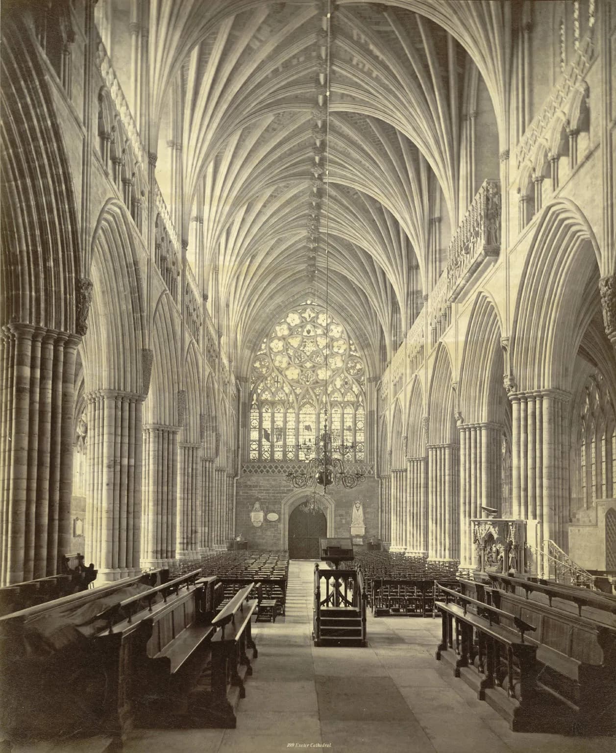 A long-perspective view of the intricately vaulted nave of a historic cathedral with rows of wooden pews and a large stained-glass window at the far end.