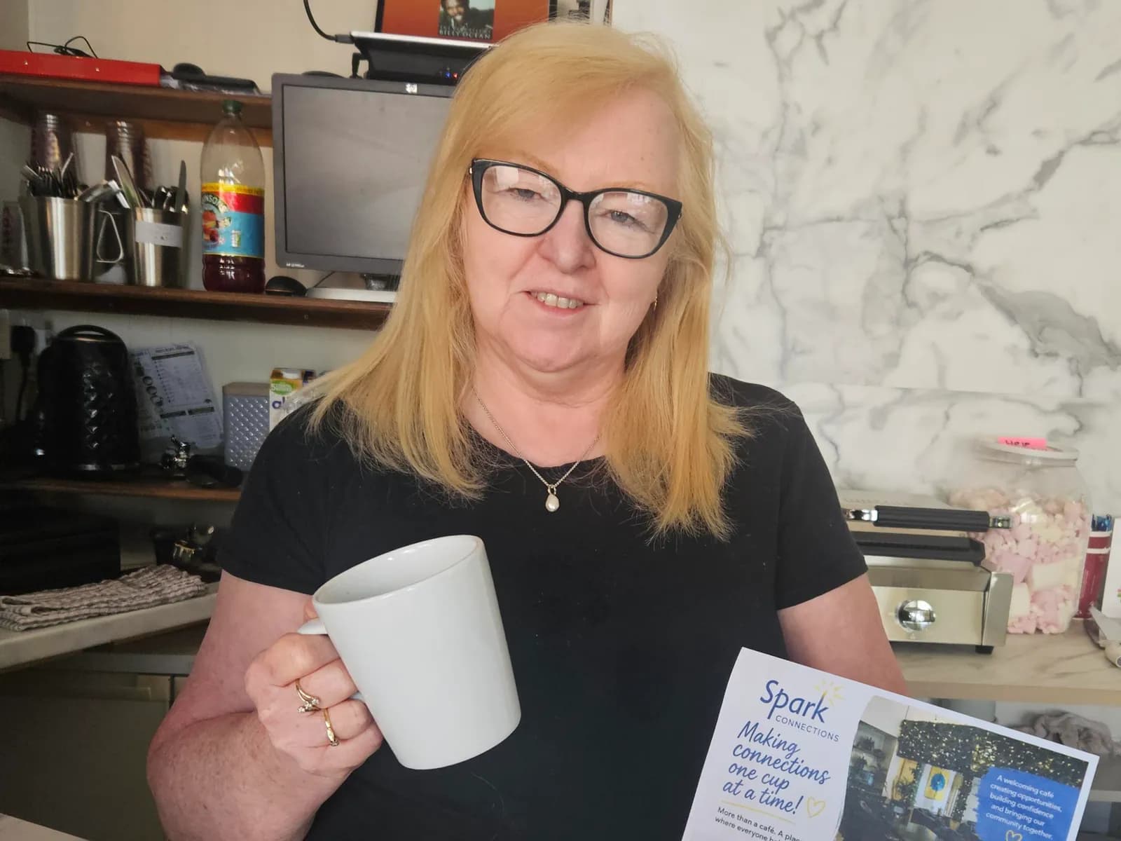 A smiling woman with blonde hair and black glasses holds a white mug and a pamphlet that reads "Spark Connections: Making connections one cup at a time!"