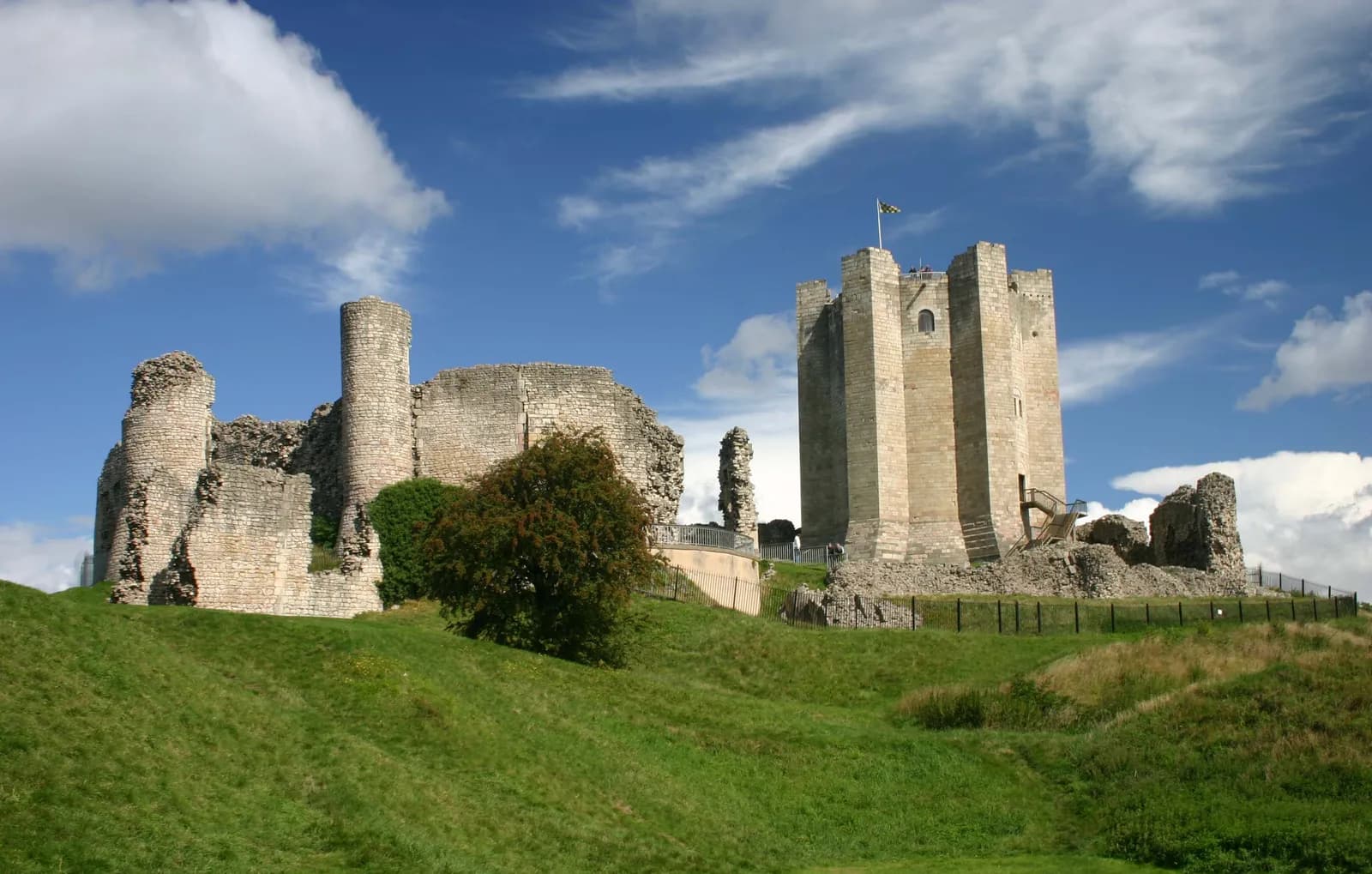 A large, partially ruined stone castle with a prominent square tower, set on a green grassy hill under a blue sky with white clouds.