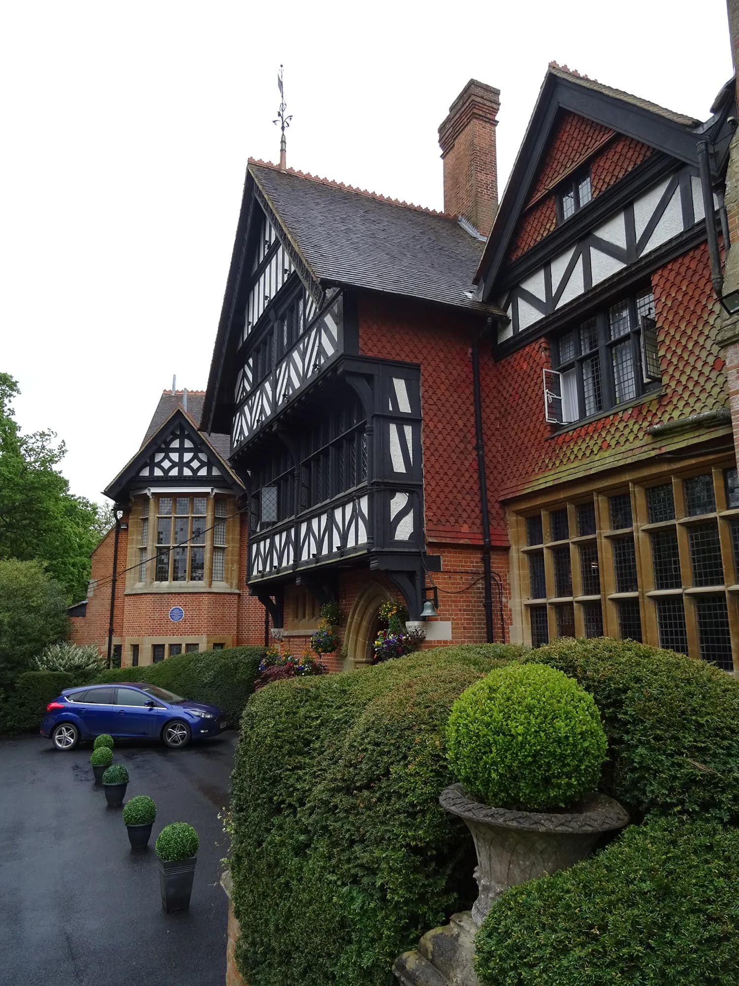 A grand, red-tiled brick house with black and white timbered gables and many windows, surrounded by sculpted green bushes and a dark driveway with a blue car parked on it.