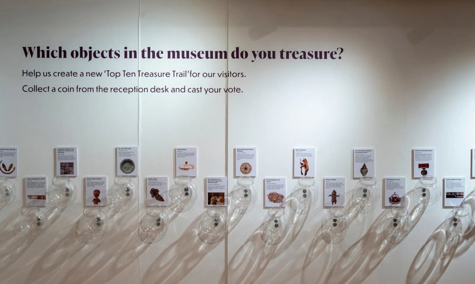 A museum exhibit wall asks visitors to choose which objects they treasure, featuring text and a series of cards displaying different artifacts with information and voting slots.