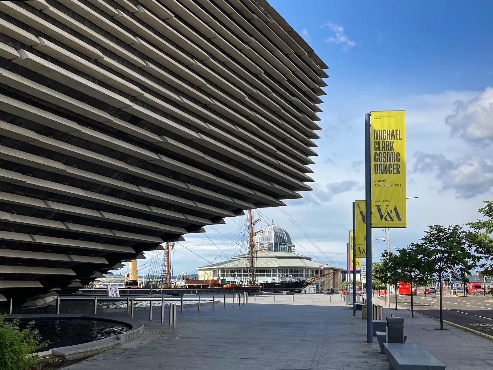 A modern building with a ribbed concrete facade next to a pathway with yellow banners advertising an exhibition, with a river and a traditional ship in the background.