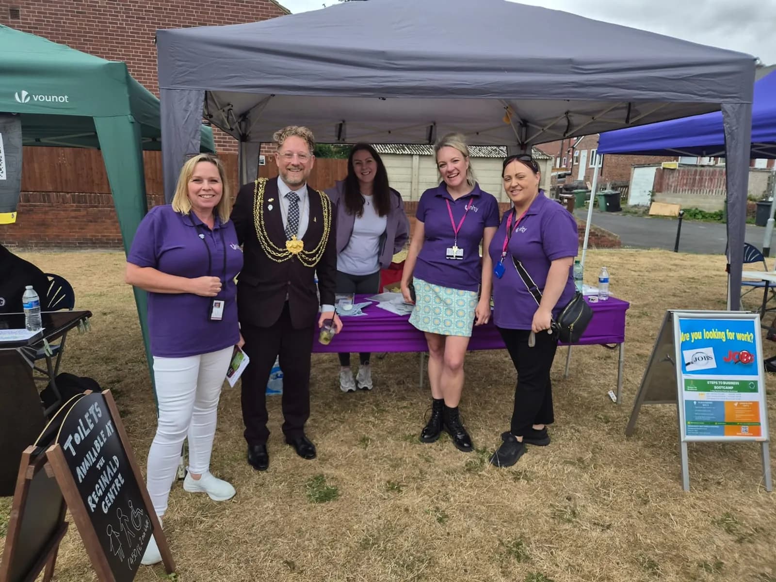 Four women and one man, all wearing purple shirts except for the man in a suit with a ceremonial chain, stand under an event tent on a grassy area.