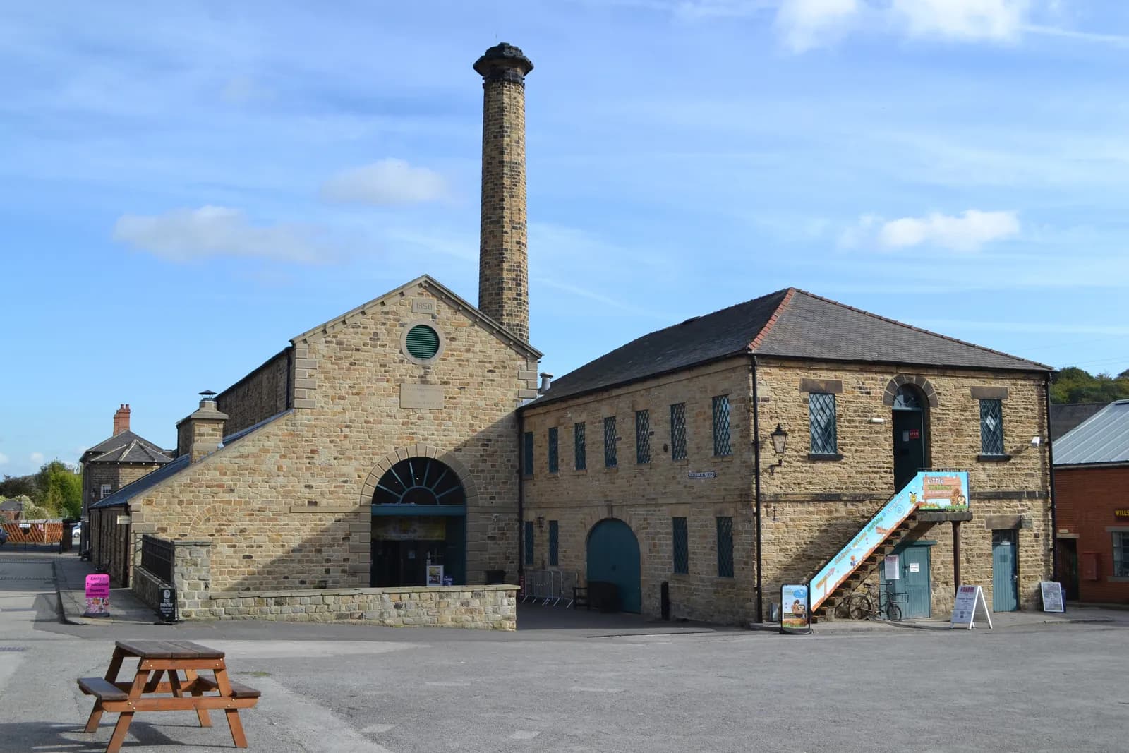 A large stone building with a tall brick chimney and an adjacent smaller stone building under a blue sky.