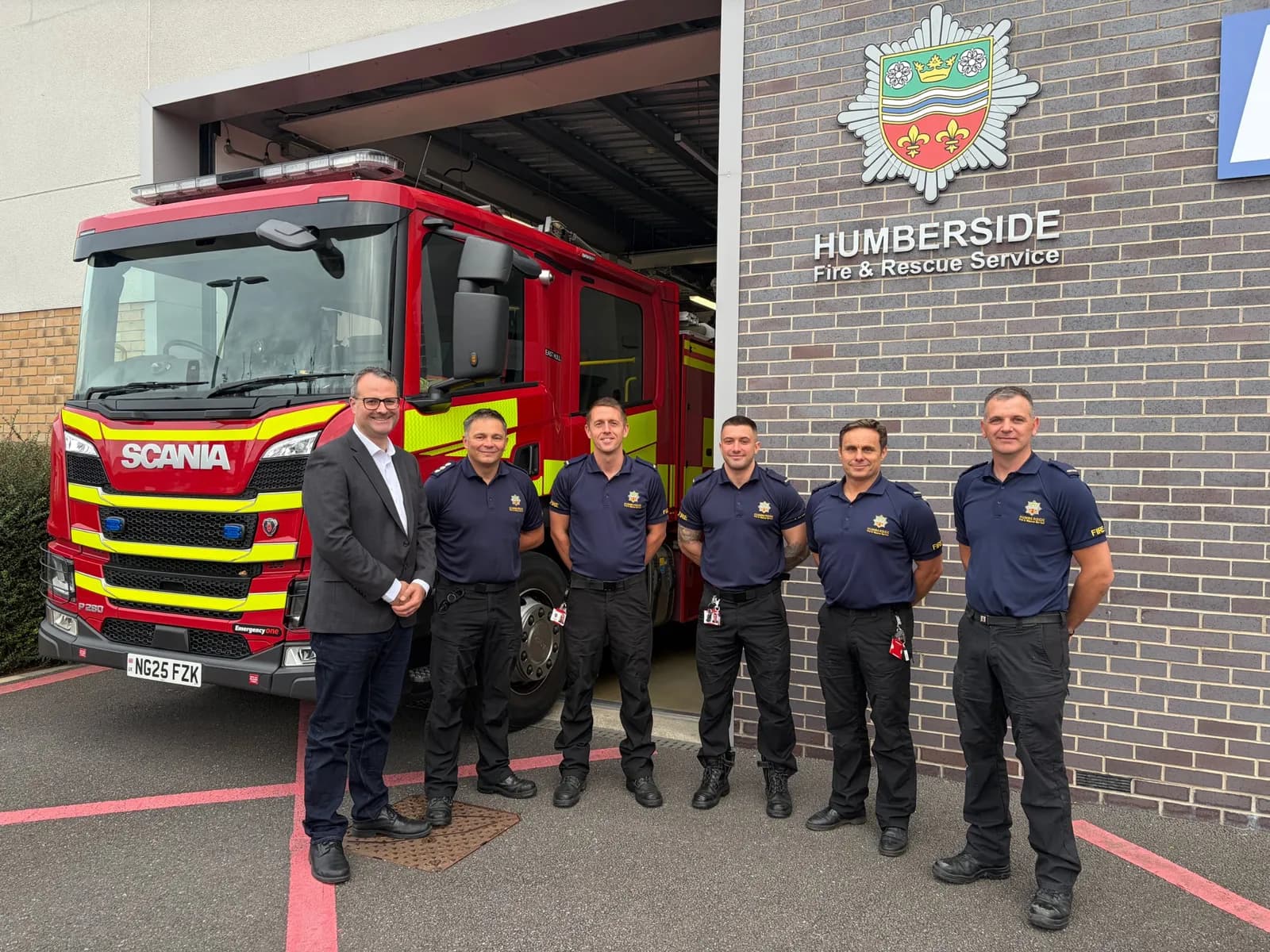 A man in a suit stands with five firefighters in navy uniforms in front of a red fire truck and a building with the Humberside Fire & Rescue Service logo.