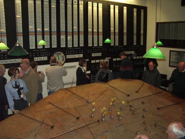 A group of people standing around a large table with a map on it while another person stands by a large display board filled with lights.