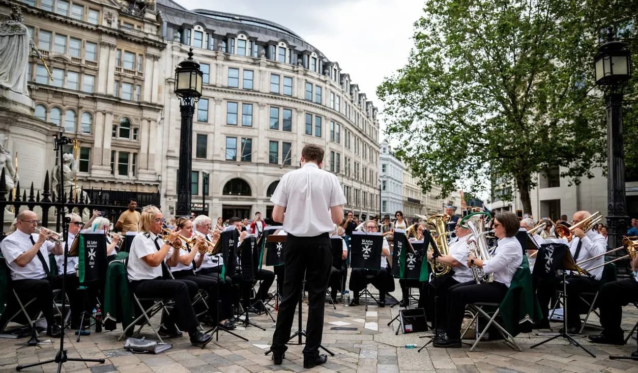 A band performs in an outdoor square surrounded by historic buildings and trees, with a conductor facing the musicians.