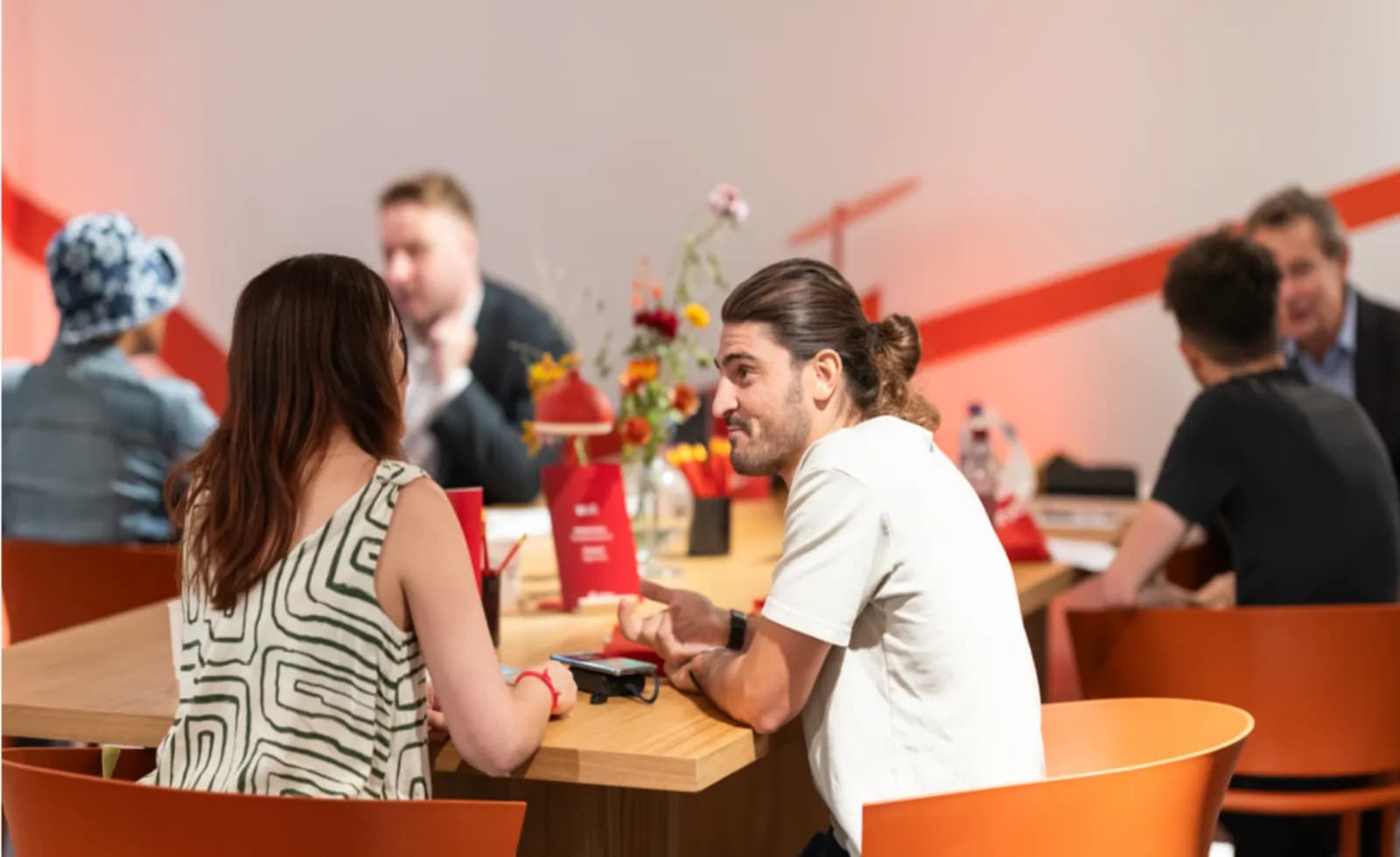 A man with long hair tied back sits at a wooden table across from a woman, both engaged in conversation with other people around the table in a brightly lit room.