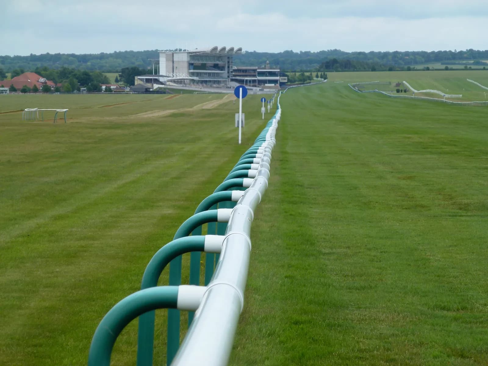 A long, curved white and green railing extends from the foreground to the midground of a green grassy field, with a grandstand building visible in the distance under a cloudy sky.