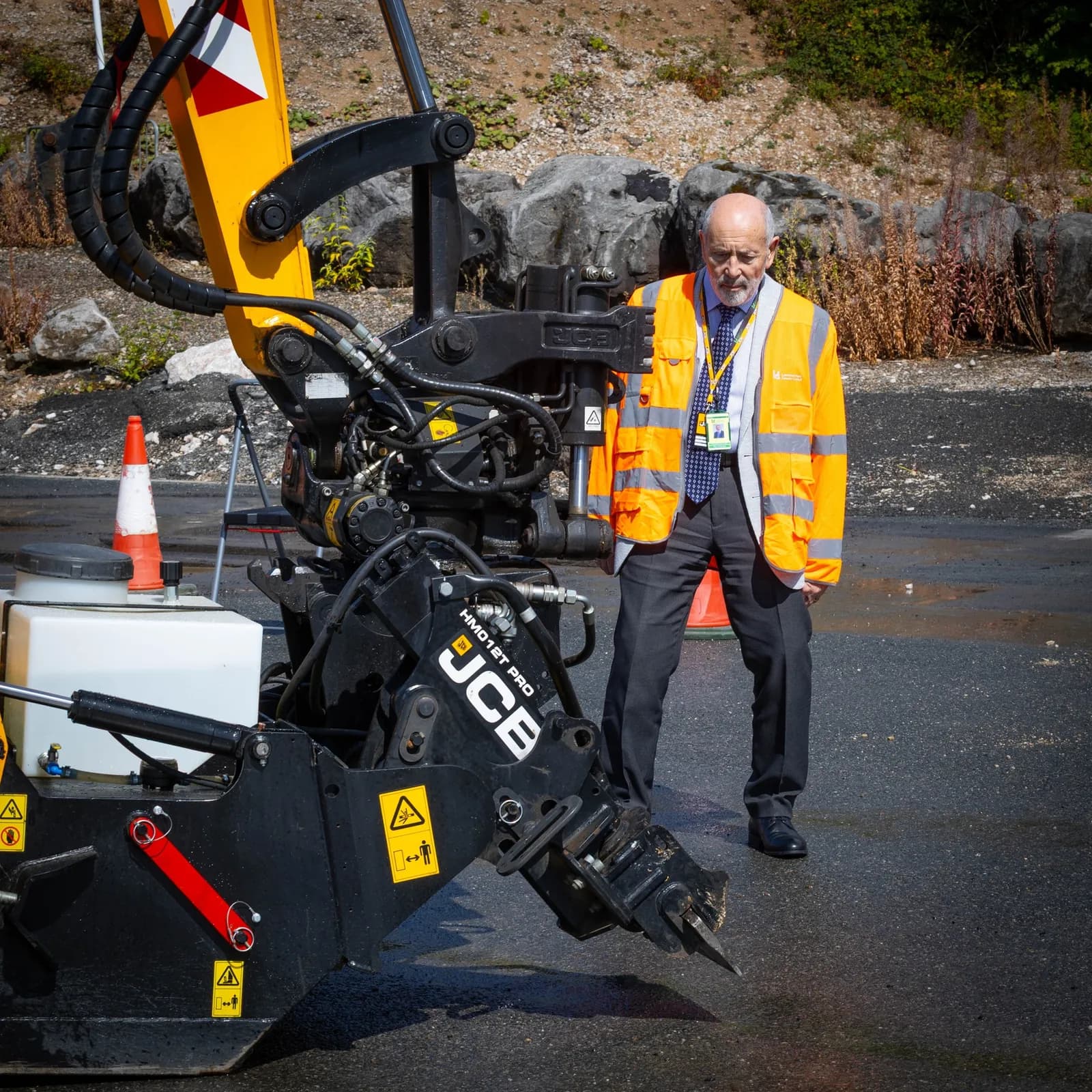 An older man in a high-visibility vest and tie stands next to the arm of a JCB excavator with a construction cone nearby.