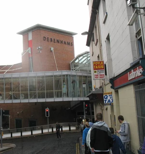A street view of a small town with a large Debenhams department store with a glass bridge leading to another building and a taxi stand in the fore, as people walk about and cross the street.
