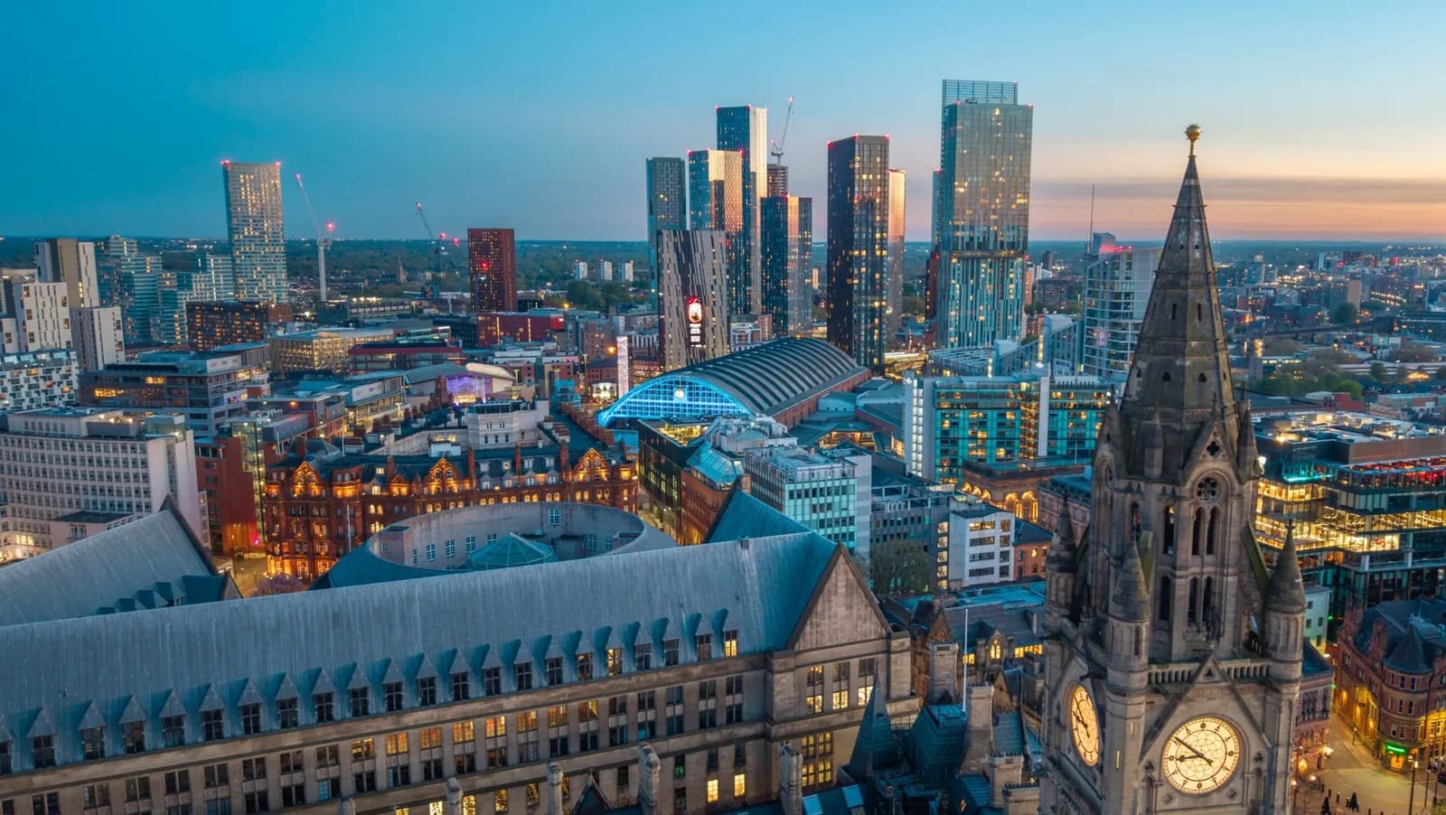An aerial view of the illuminated Manchester cityscape at dusk, featuring the clock tower of Manchester Town Hall in the foreground against a backdrop of modern skyscrapers and other city buildings.