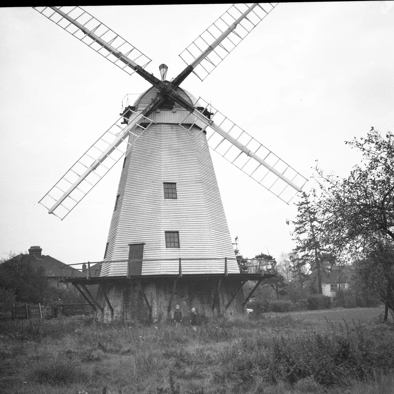 A black and white photograph shows a tall, hexagonal windmill with four large sails, nestled in a grassy field on an overcast day.