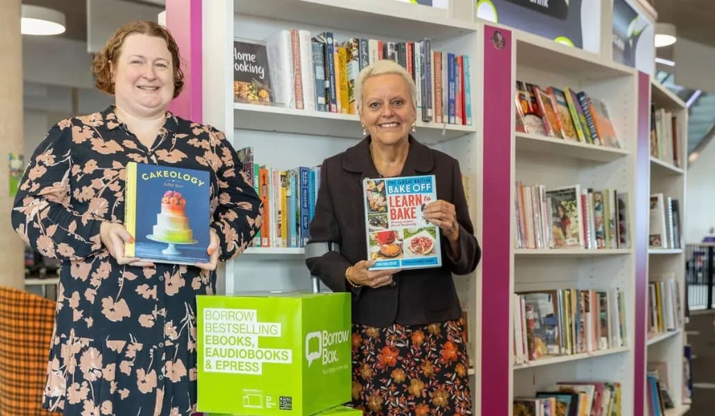 Two women stand in a library, each holding a baking cookbook, with green "Borrow Box" containers visible below them.