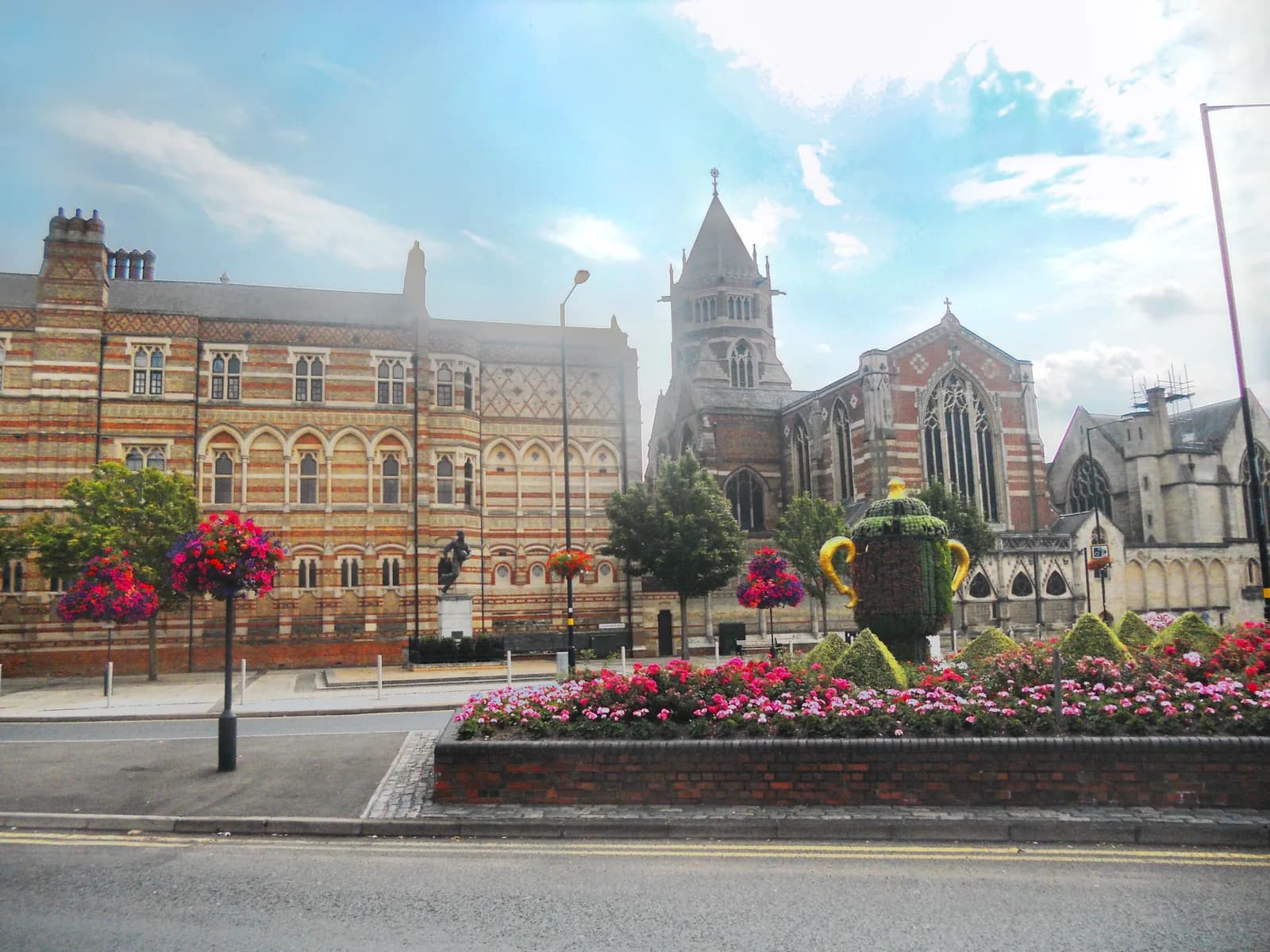 A large building with brick and concrete patterns on its facade, a tall church with a prominent steeple, and a decorative garden with a teapot-shaped topiary and colorful flowers in the foreground.