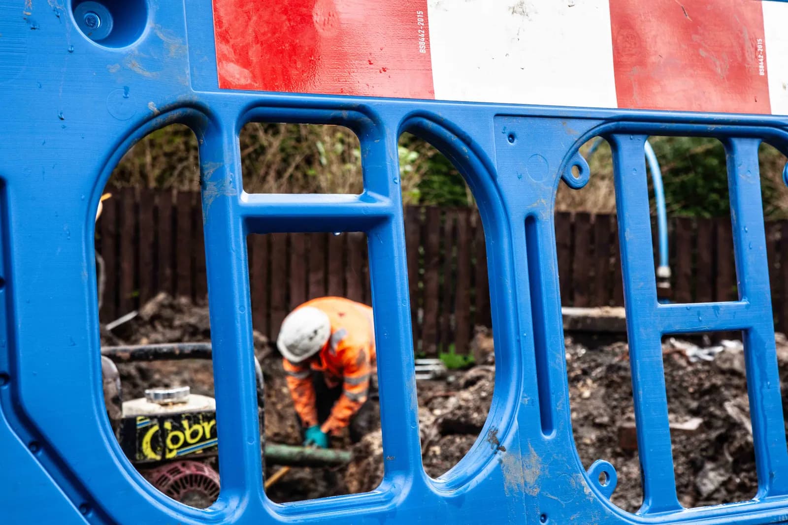 A construction worker in a hard hat and orange high-visibility jacket is seen through blue safety barriers, bent over working in a ditch.