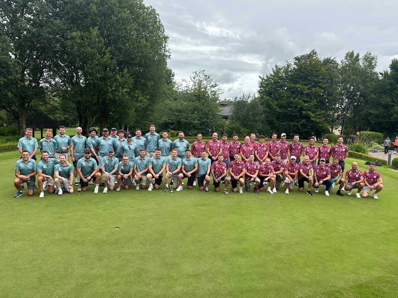 A large group of men, some standing and some kneeling, pose for a photo on a golf course with trees and a cloudy sky in the background.