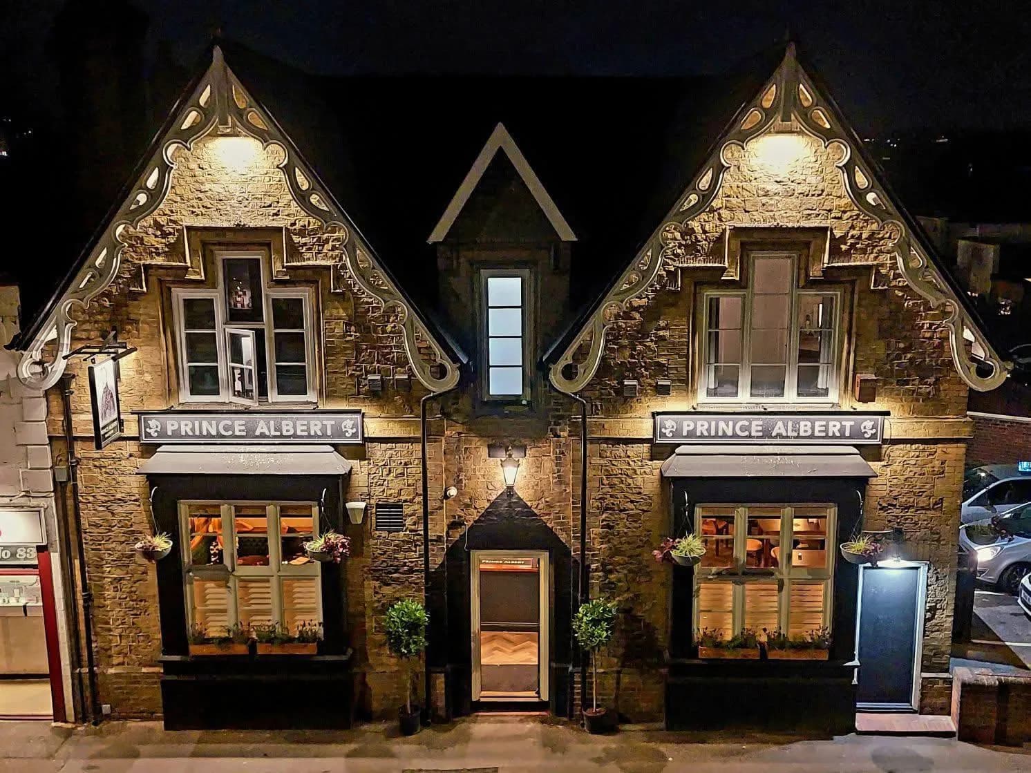 A two-story building with a dark roof and illuminated cream-colored stone walls at night, featuring a sign that reads "Prince Albert."