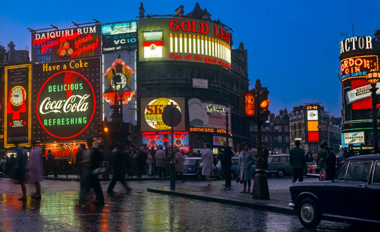 A nighttime street scene displays large, vibrant neon advertisements illuminating a building and the wet street below, with blurred figures of people and cars moving.