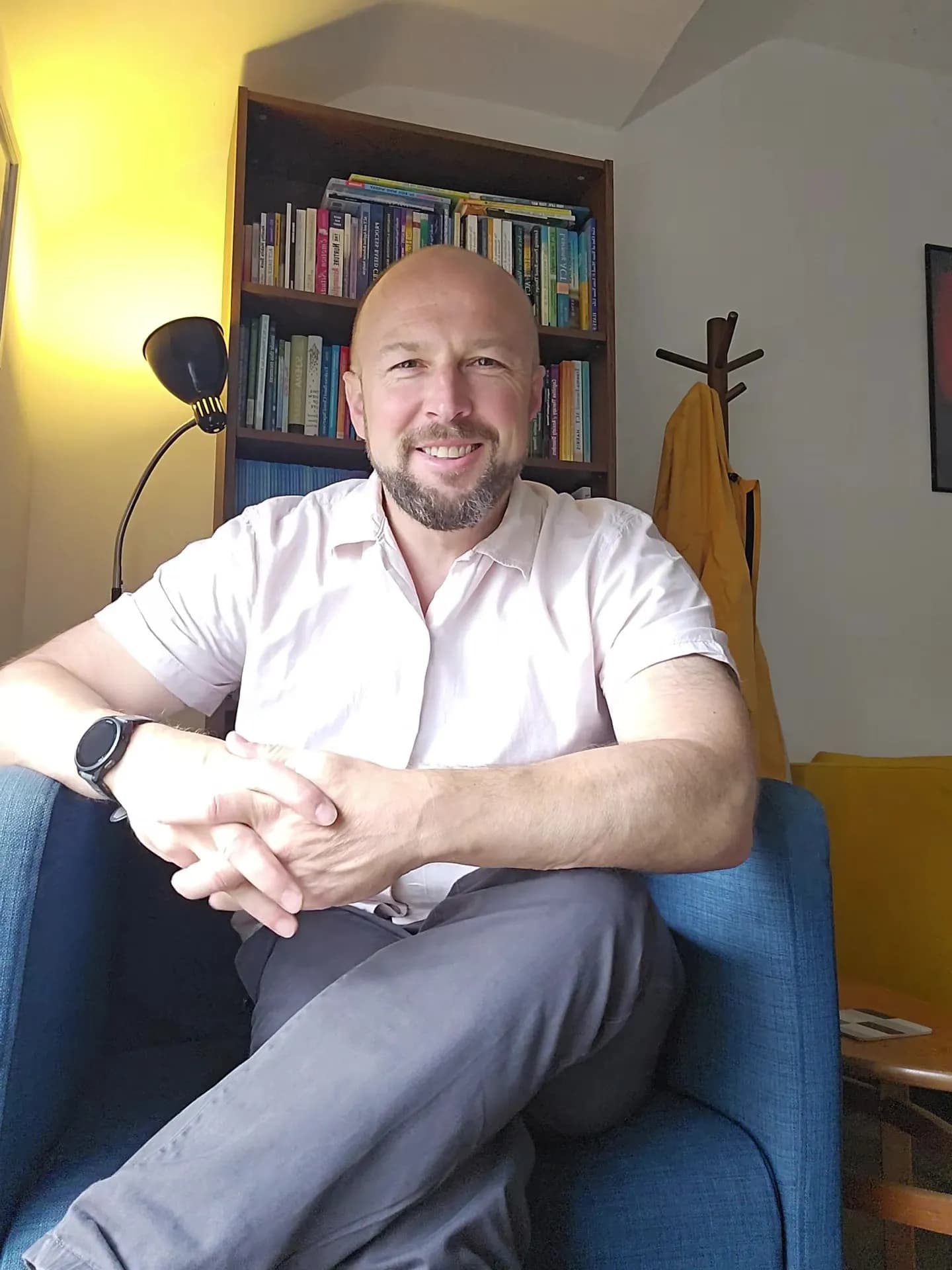 A smiling man with a beard and bald head wearing a light-colored shirt and grey trousers sits in a blue armchair in a room with a bookshelf behind him.