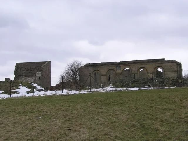 A cloudy day shows the ruins of a stone building with arched windows, while snow and sparse trees dot the landscape behind a fence and a field of green and brown grass in the foreground.