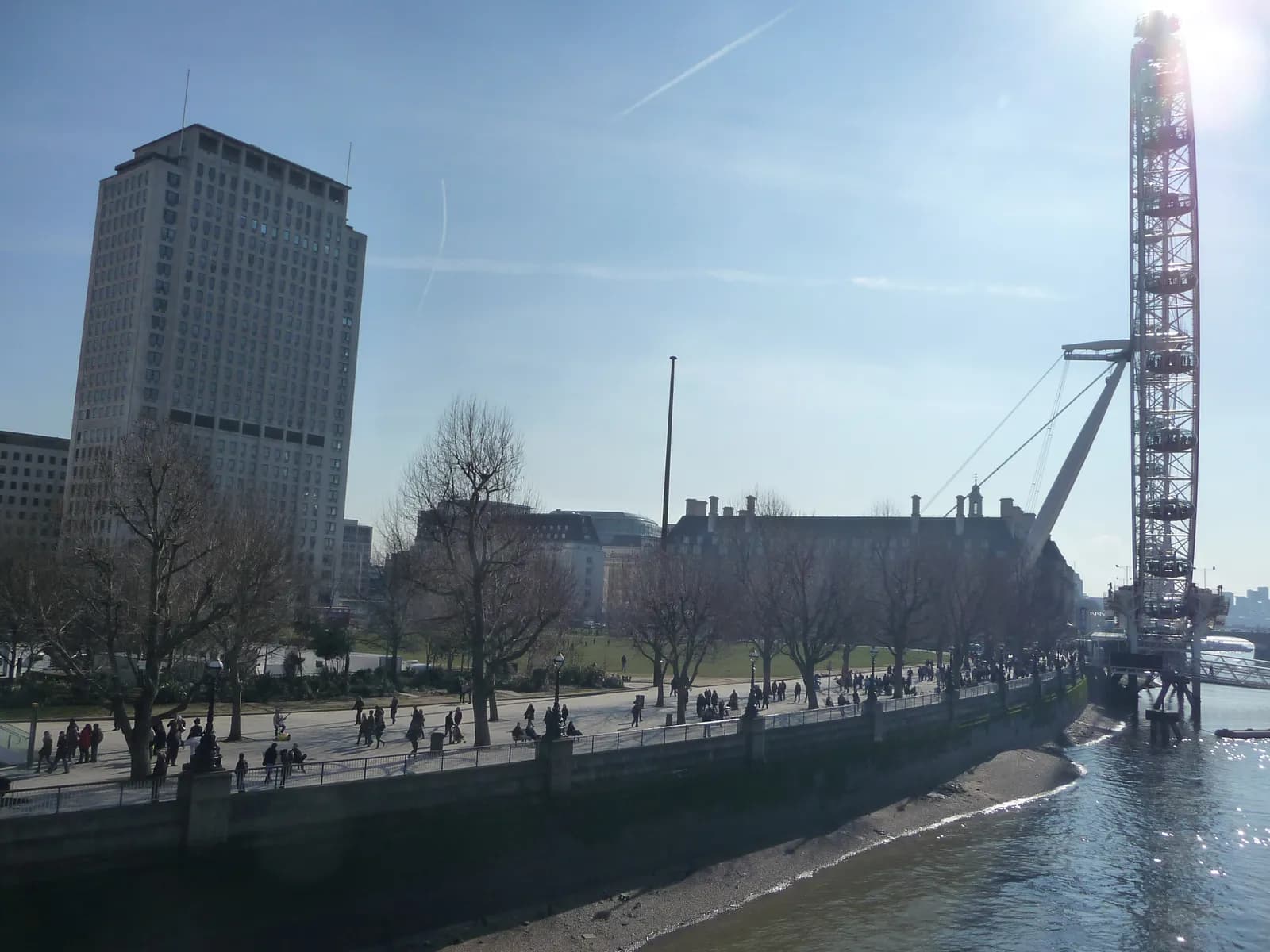 A wide shot shows a tall building on the left and the lower half of the London Eye on the right, with a river bank and walking path filled with people in between.