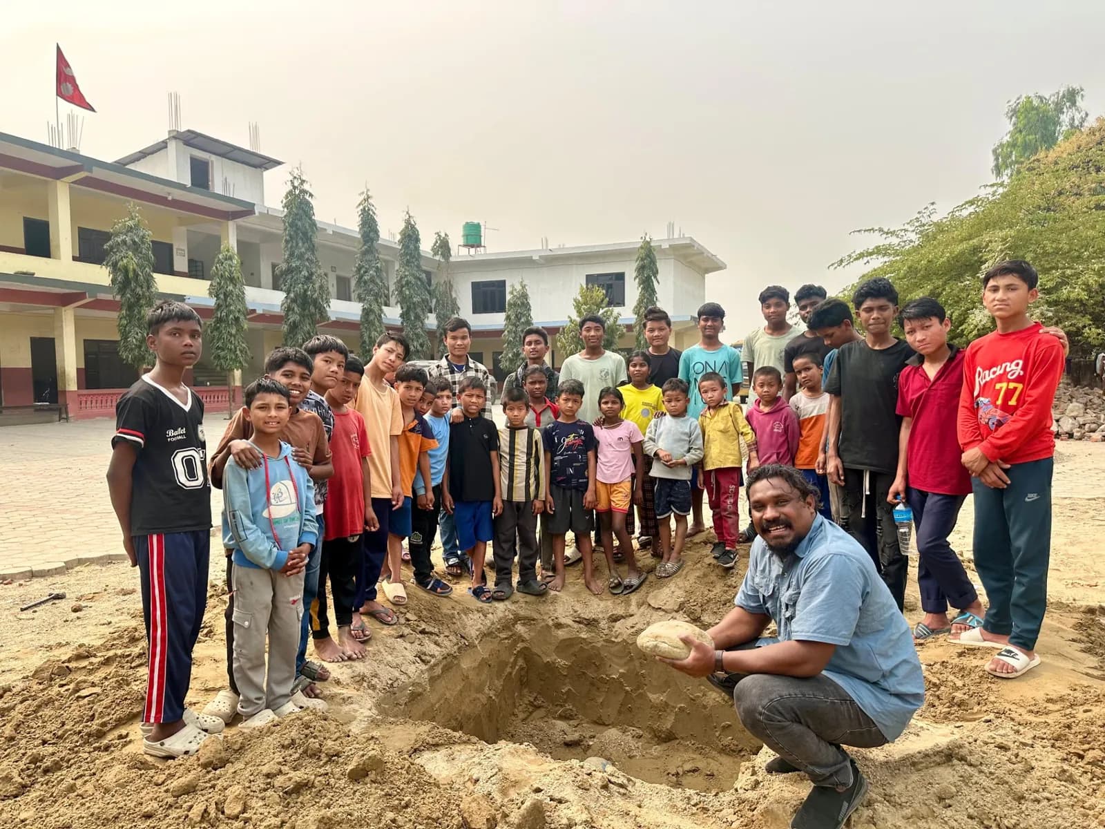 A group of children watch a man squatting with a rock in a dug out pit, with a school building in the background.