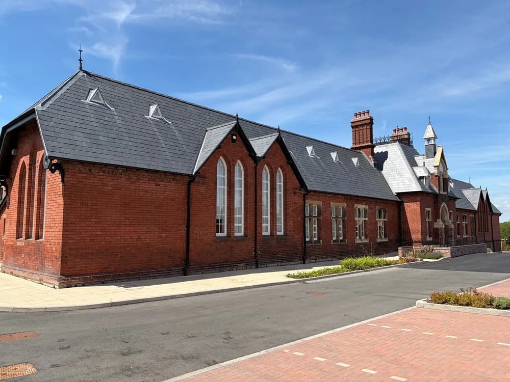 A long brick building with a dark gray tiled roof, multiple tall arched windows, and chimneys, sits alongside a paved road under a blue sky.
