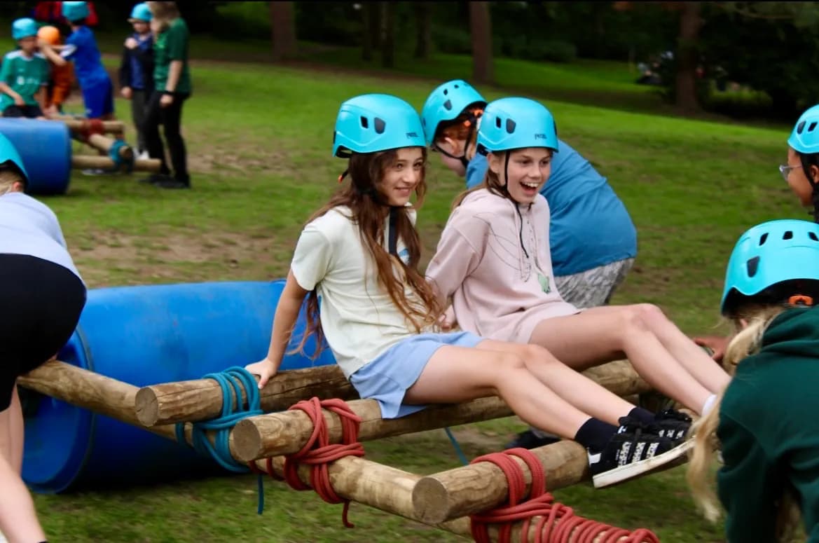 Two smiling girls wearing blue helmets sit on a wooden obstacle course, surrounded by other children and green grass.