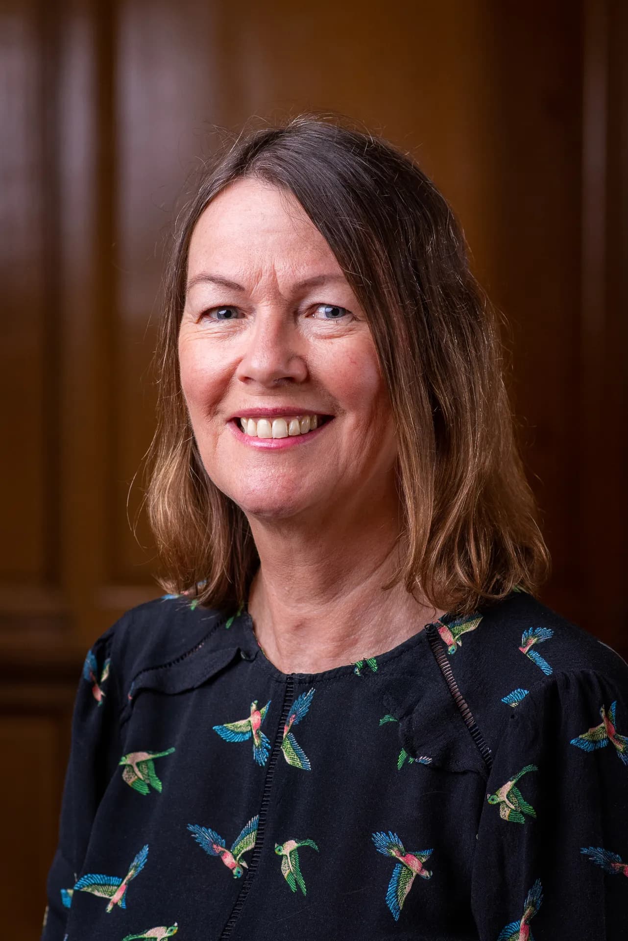 A woman with light brown hair and a dark shirt with colorful bird patterns smiles at the camera against a dark wooden background.