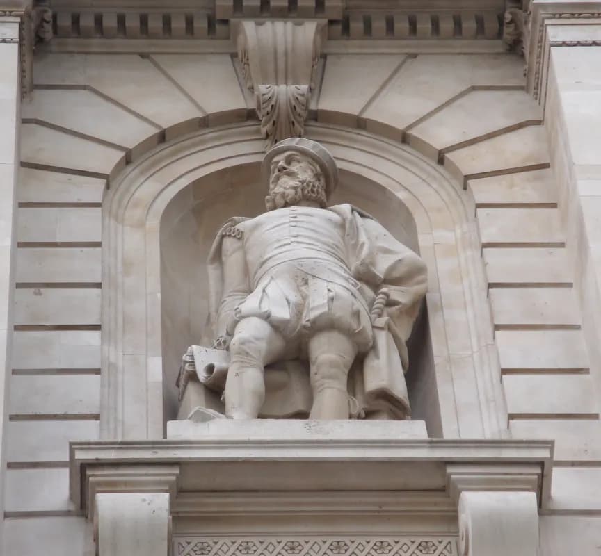 A light-colored stone statue of a bearded man in historical clothing stands within an arched niche on a building facade.